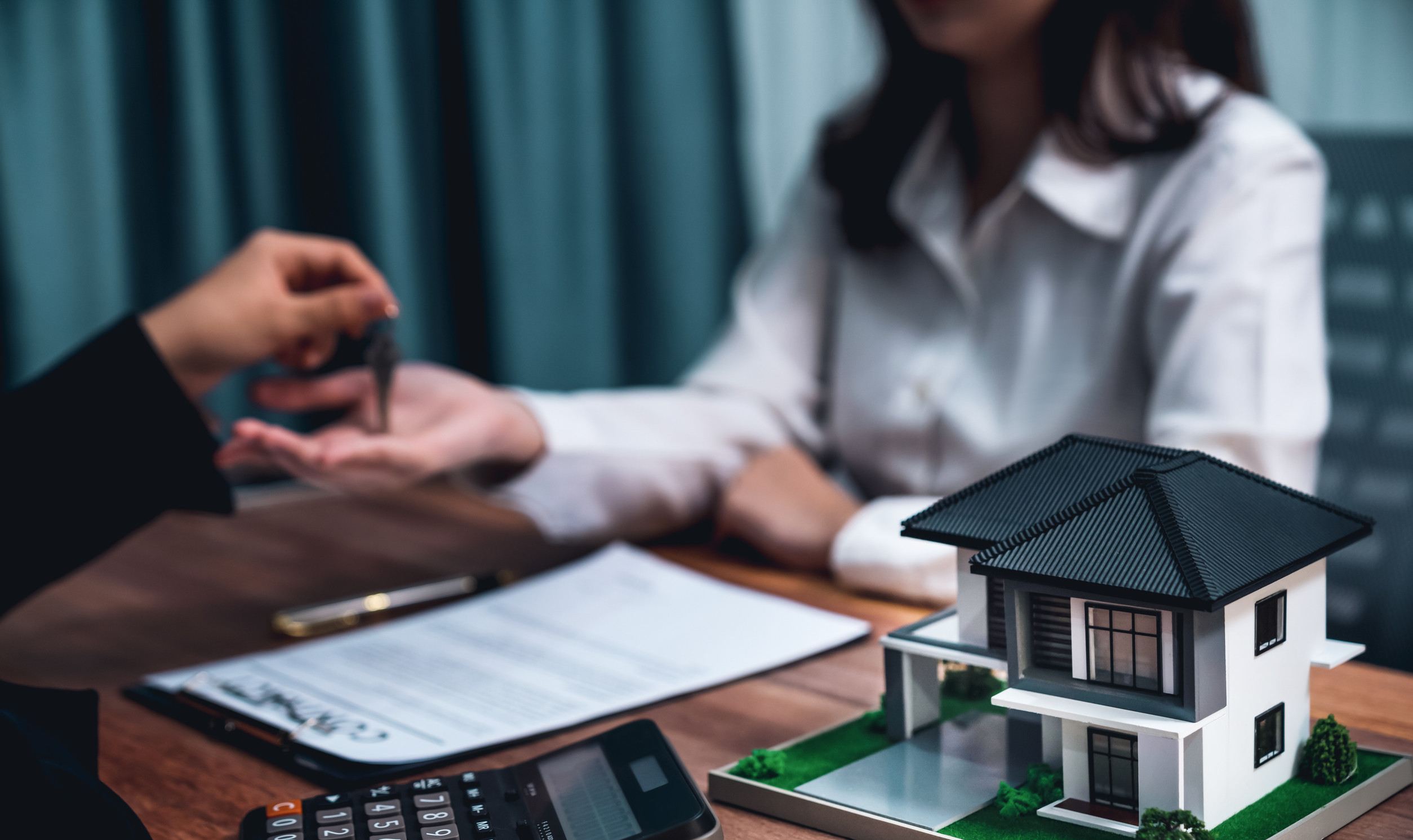 Focus house model on blur real estate agent and buyer background. Enthusiastic Person handing over keys to another individual with a model house and mortgage documents on a table, symbolizing home buying and mortgage services in Victoria.