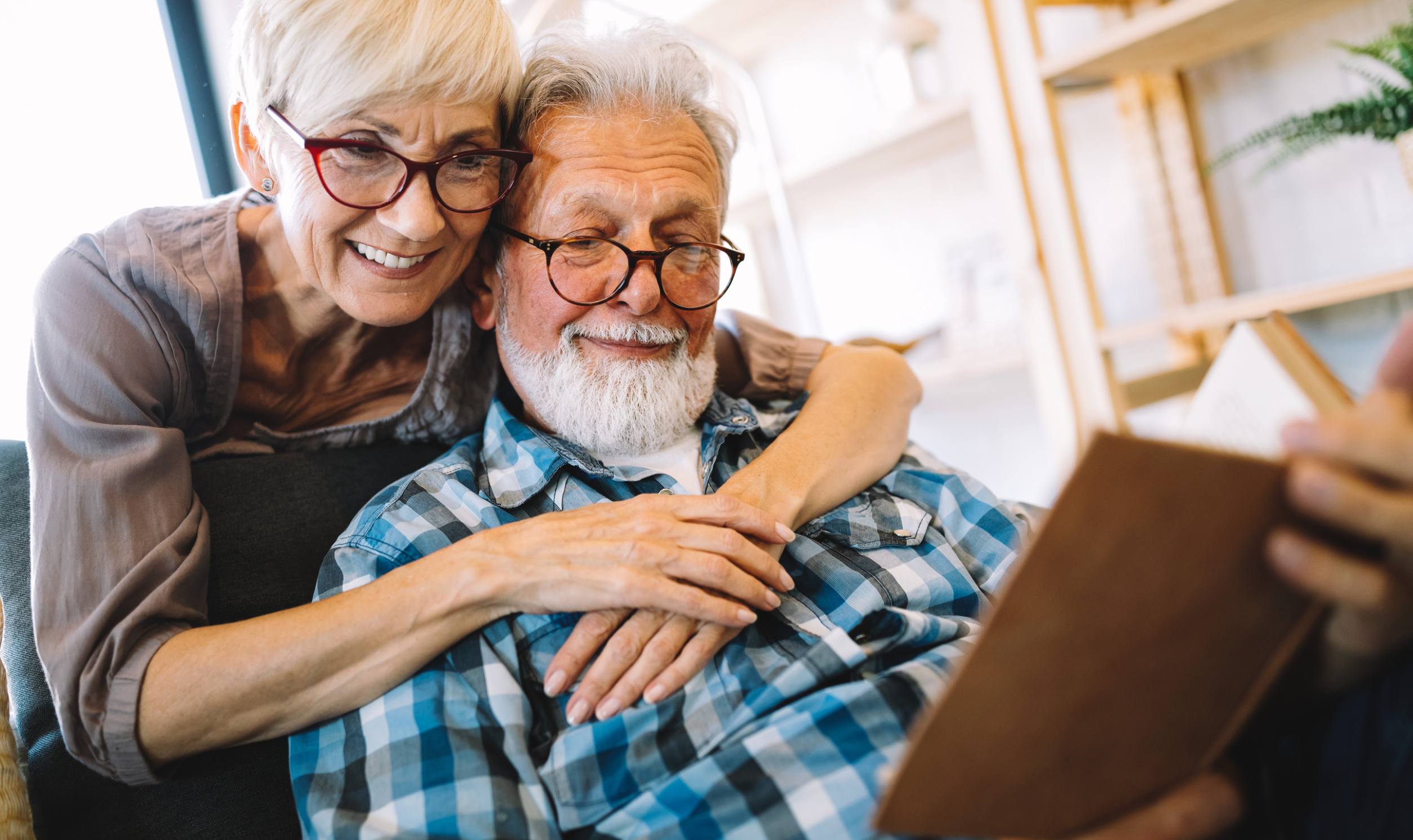 Focus house model on blur real estate agent and buyer background. Enthusiastic Elderly couple enjoying time together, with woman smiling and embracing man reading a book, reflecting warmth and companionship in a cozy home setting.