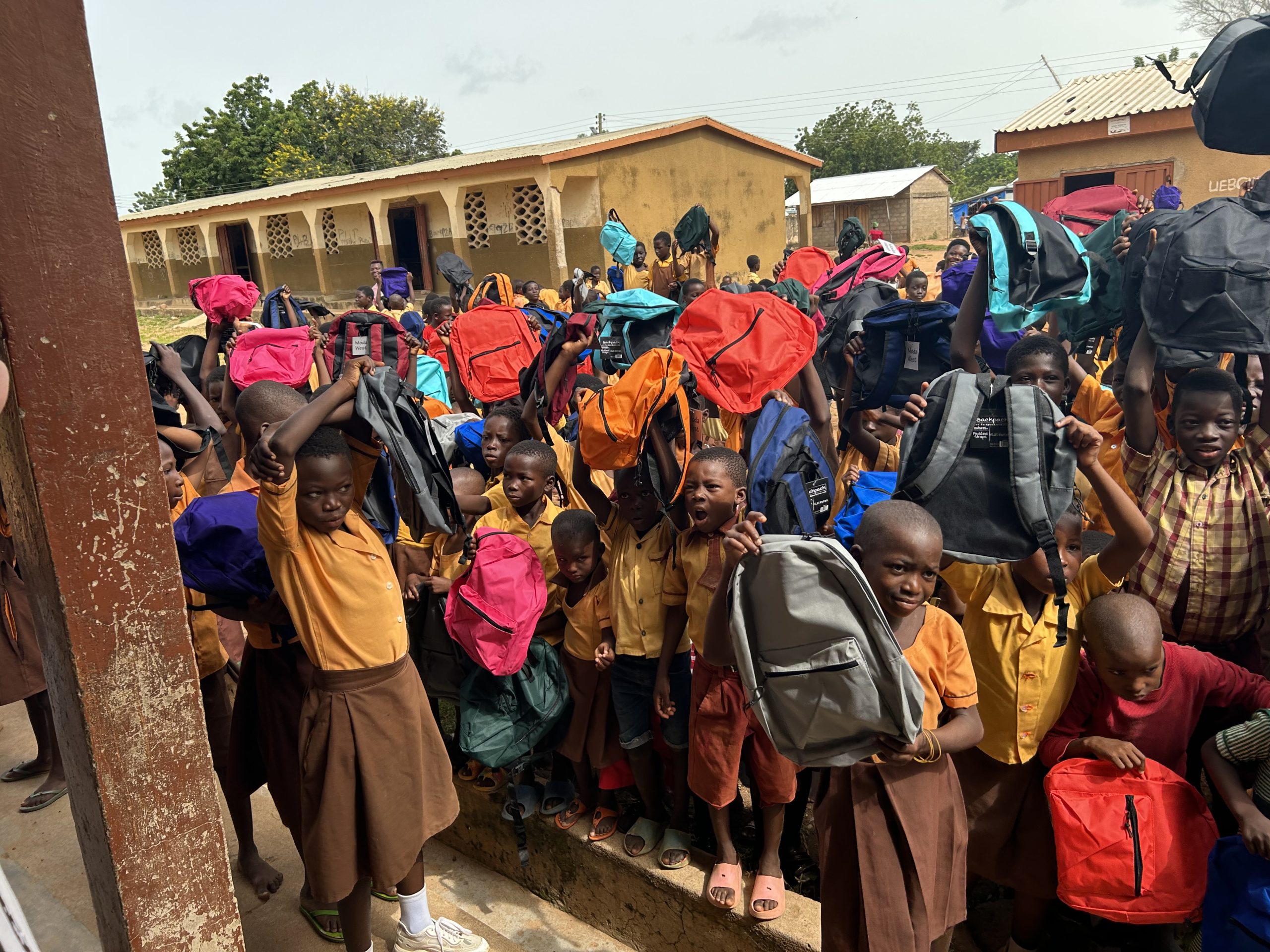 Traci Green Ghana Children in school uniforms joyfully holding colorful backpacks during a donation event in Vea, Ghana, supported by Talata’s Project.
